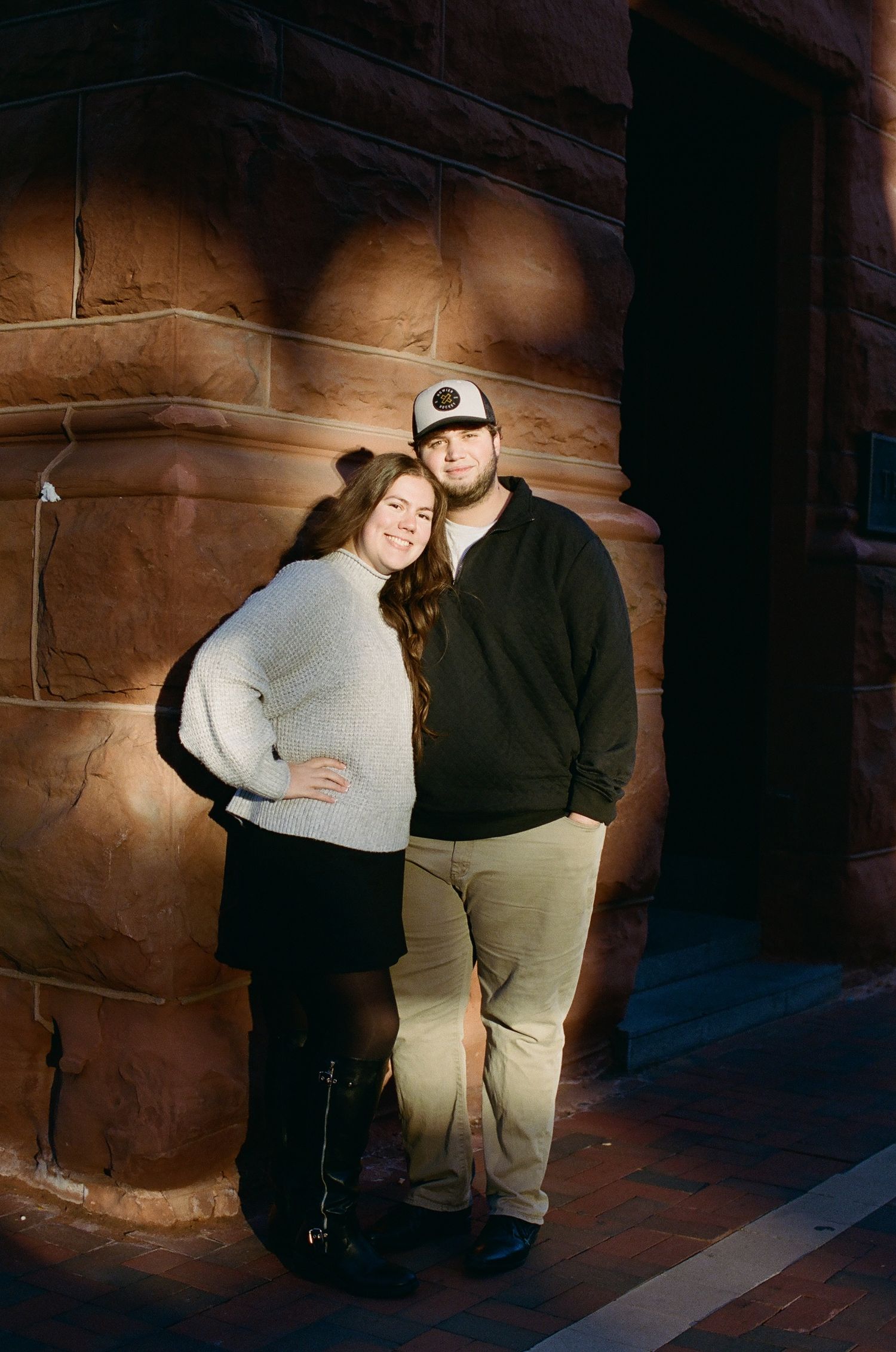 A couple poses together against a rustic stone wall in warm lighting.
