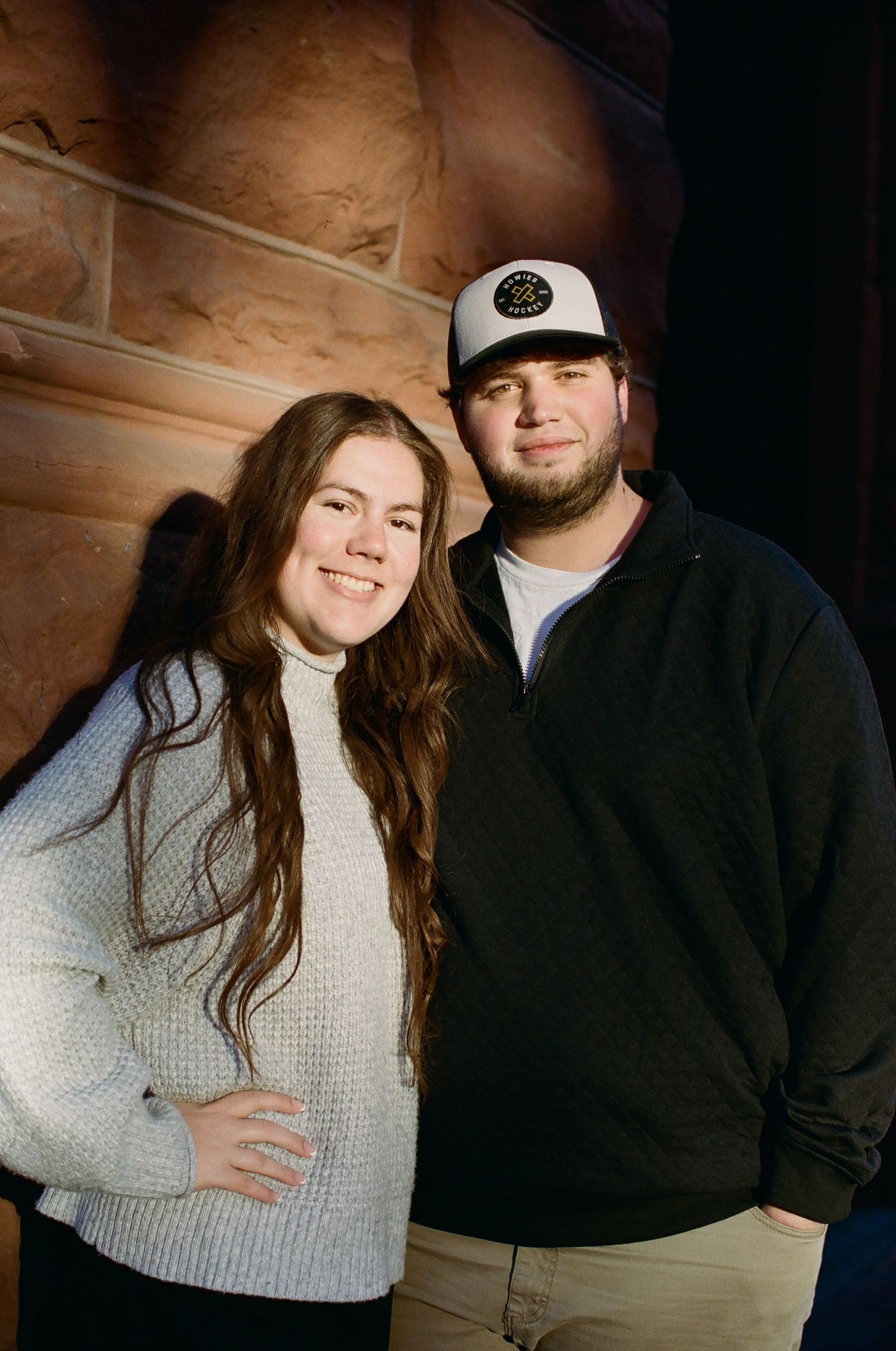 A couple poses together in a rustic setting wearing casual winter attire with a cream sweater and dark jacket.