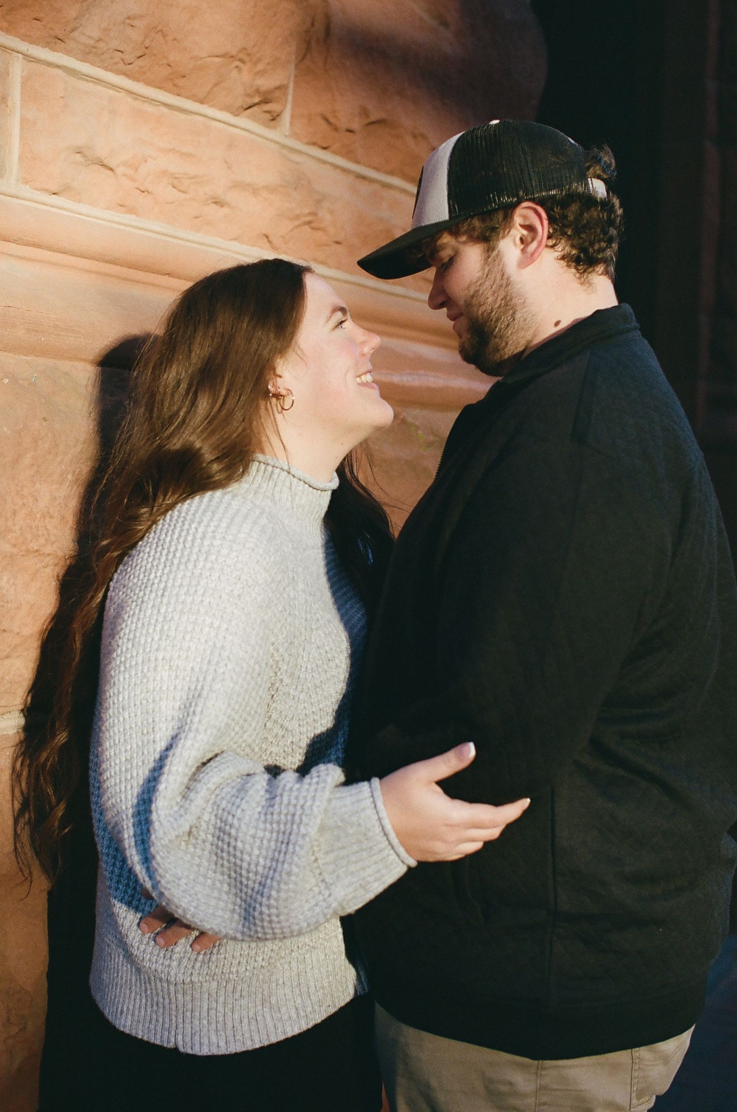 A couple shares an intimate moment against a wooden wall, with her wearing a cream sweater and him in dark clothing.