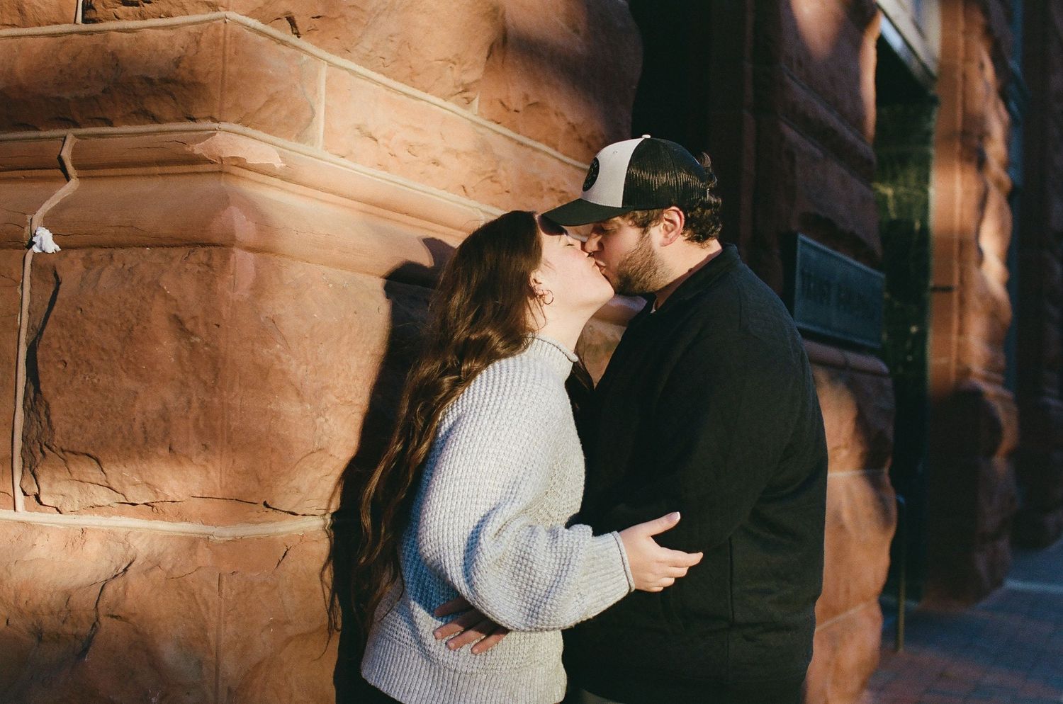 A couple embraces in a romantic moment against a rustic stone wall with warm lighting.
