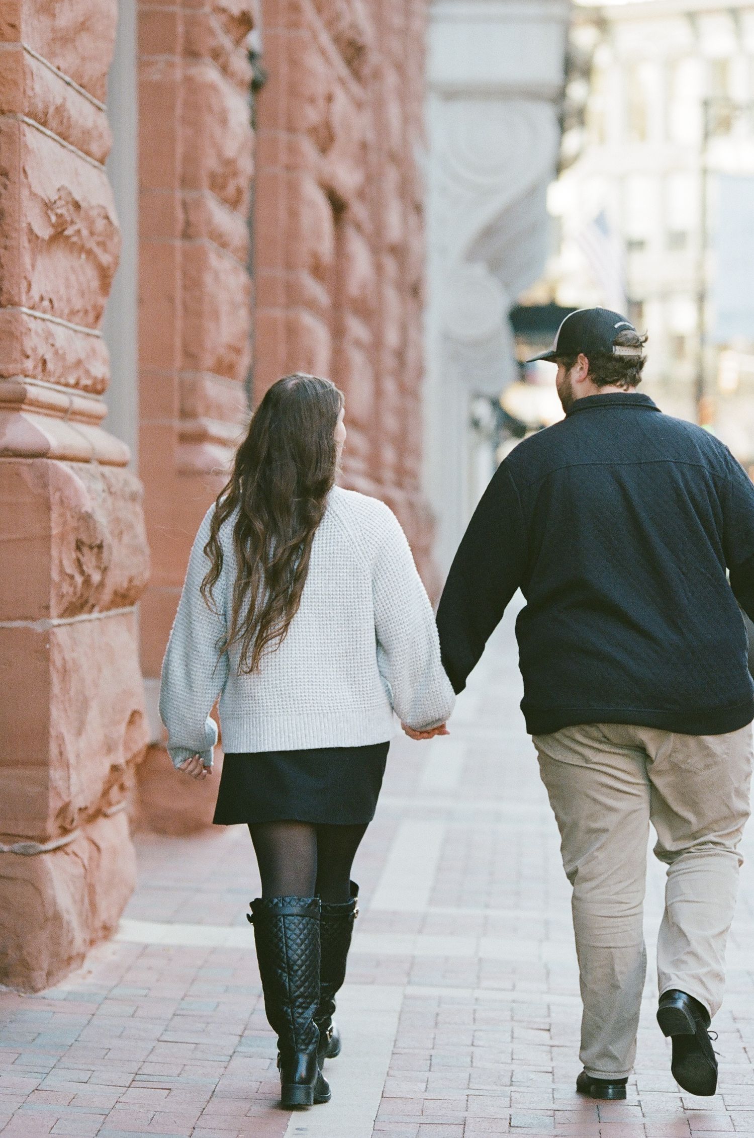 A couple walking hand in hand down a brick-lined walkway with ornate architectural details.
