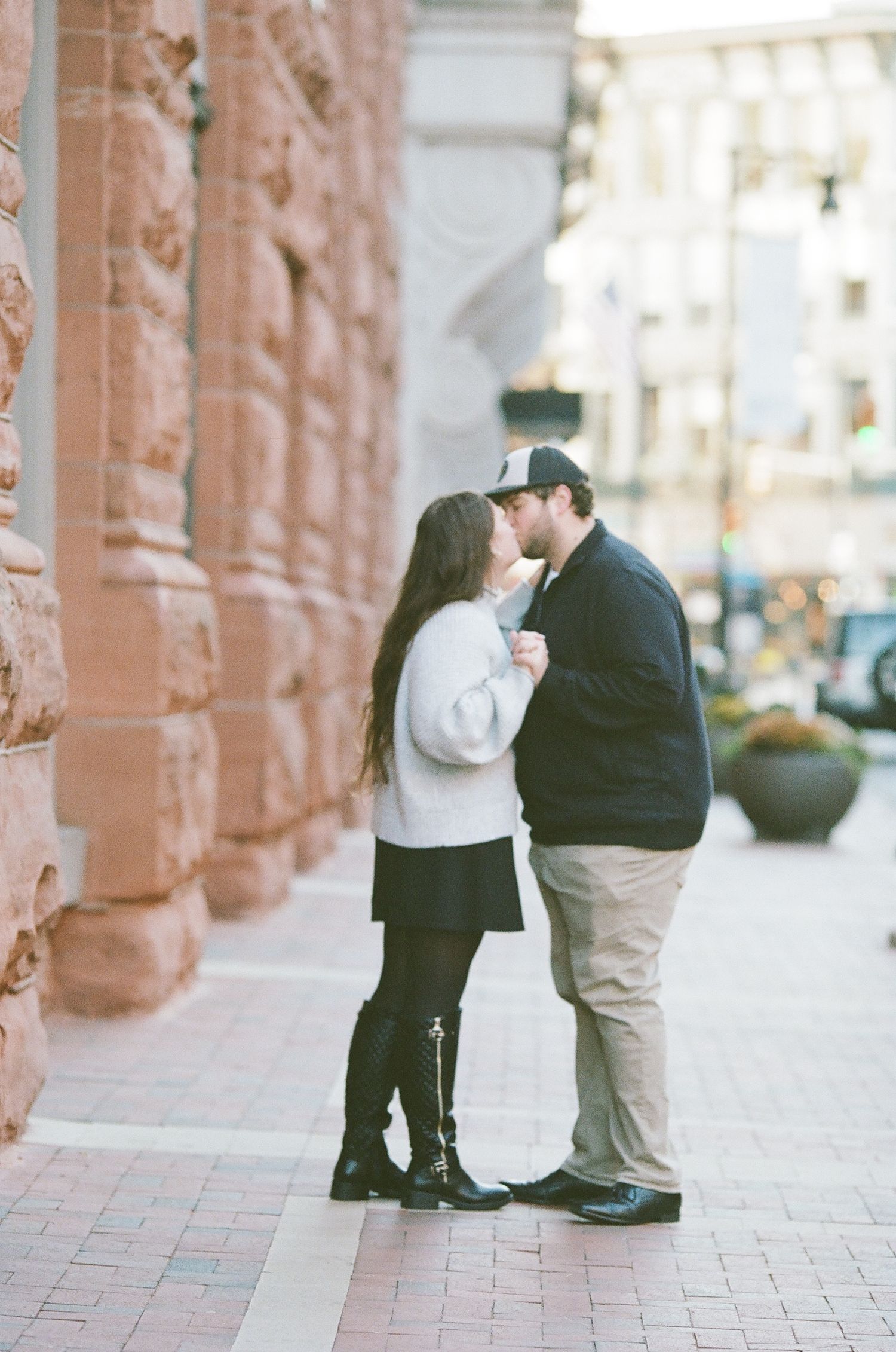 A couple shares an intimate moment under ornate stone archways along a city sidewalk.