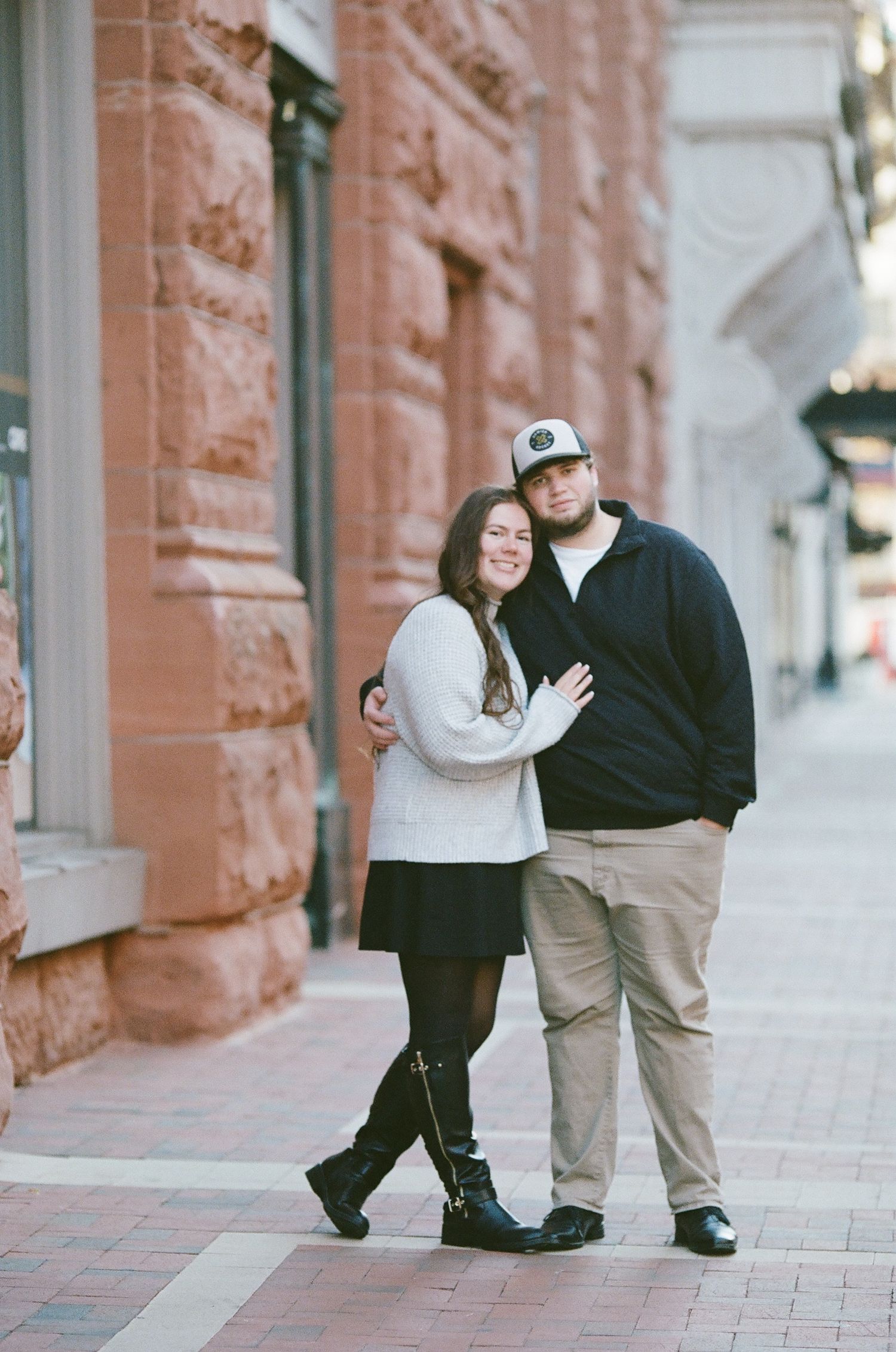 A couple shares a tender moment together on a brick sidewalk outside a historic building.
