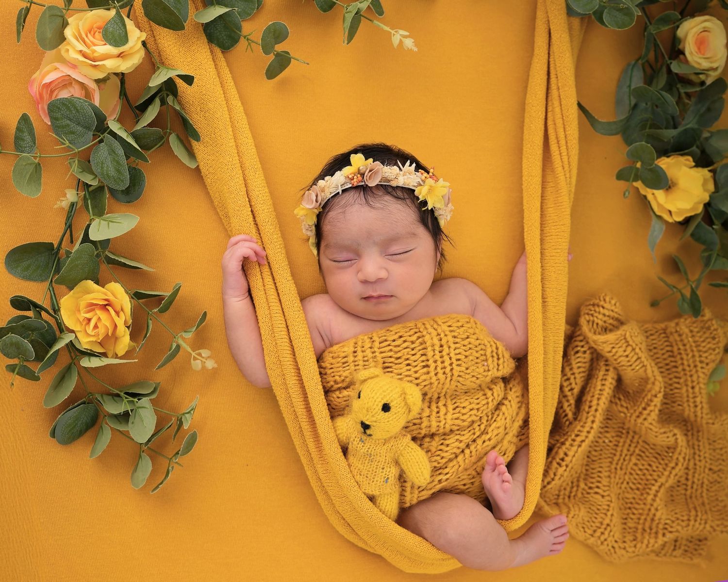 Sleeping newborn wearing yellow crown rests peacefully on golden fabric with green floral decorations.