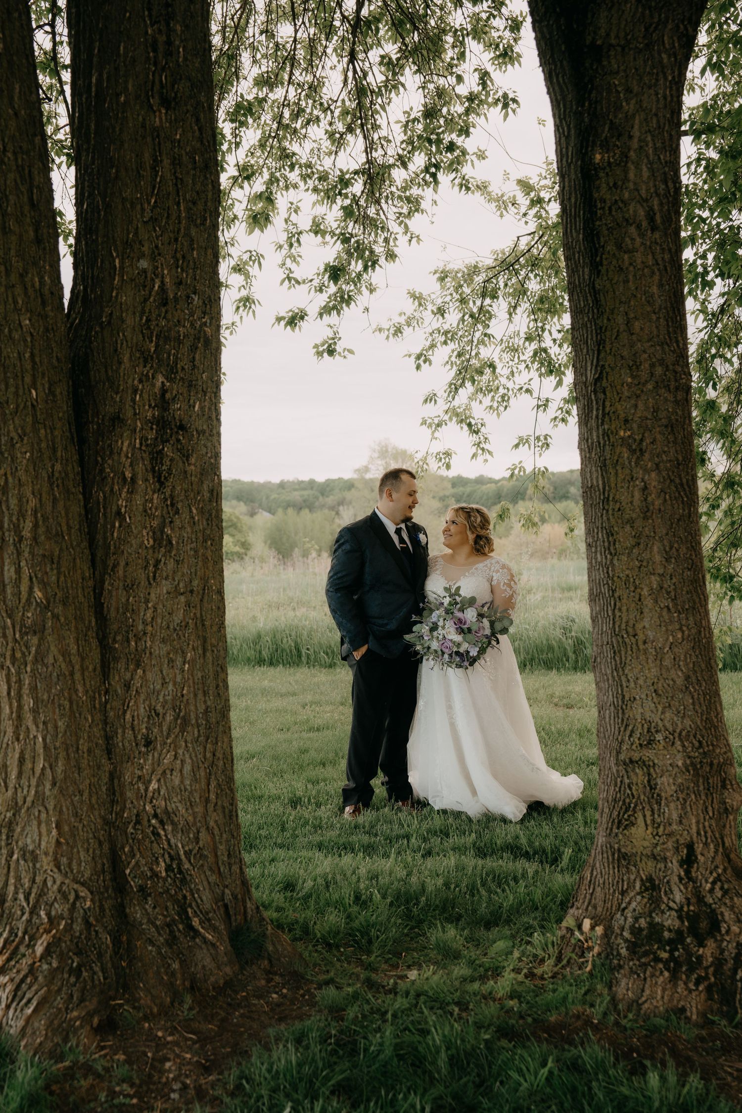 A couple shares an intimate moment between large tree trunks in a rustic outdoor wedding setting.