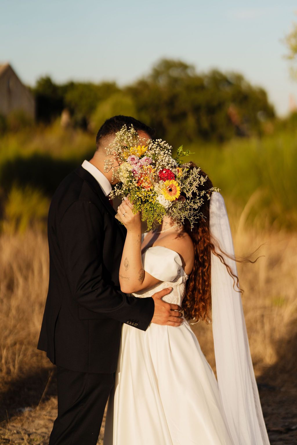 A couple shares a romantic moment in a golden field at sunset, with the bride holding a colorful wildflower bouquet.