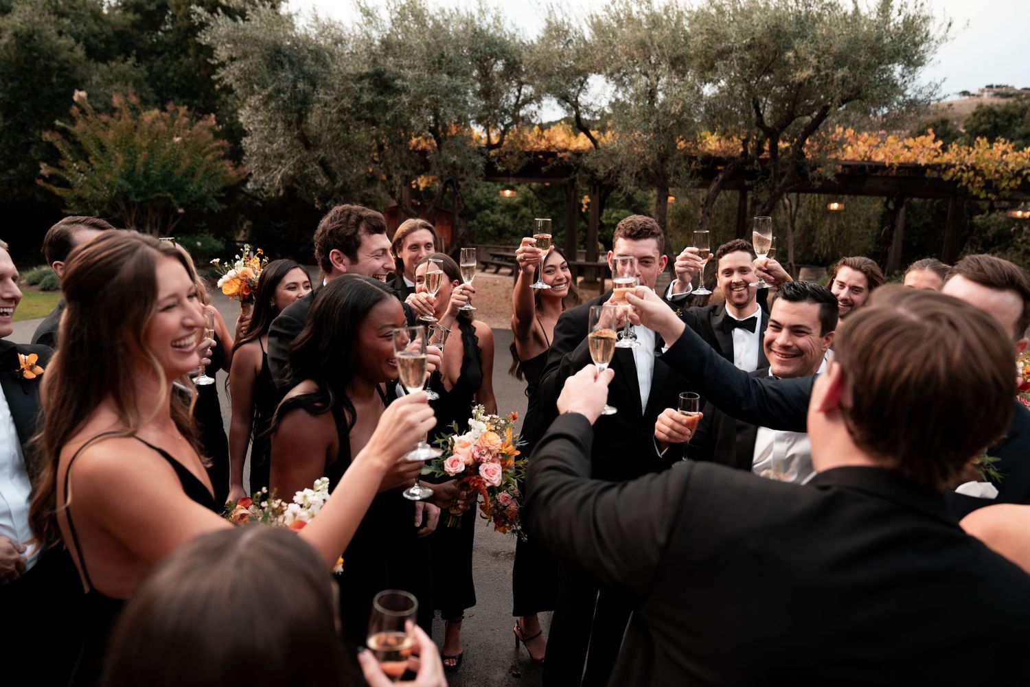 A group of wedding guests celebrate and dance together in front of a rustic stone wall during an outdoor reception.
