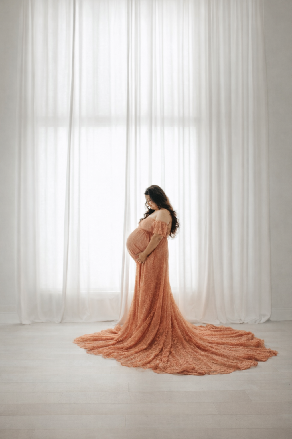 A flowing peach-colored dress creates a dramatic silhouette against white curtains in a maternity photo session.