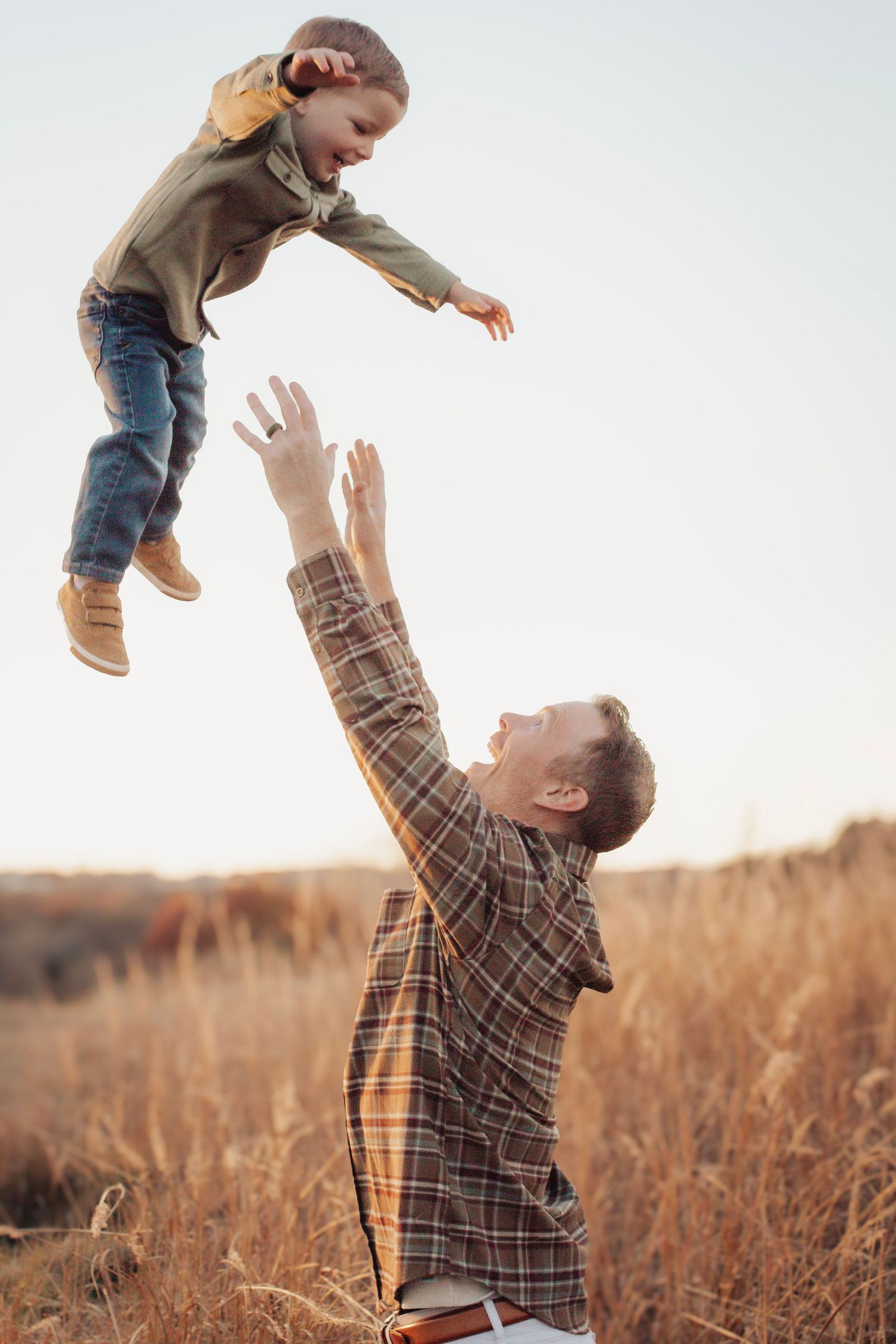 A parent playfully lifts and tosses a small child in the air while standing in a golden wheat field at sunset.