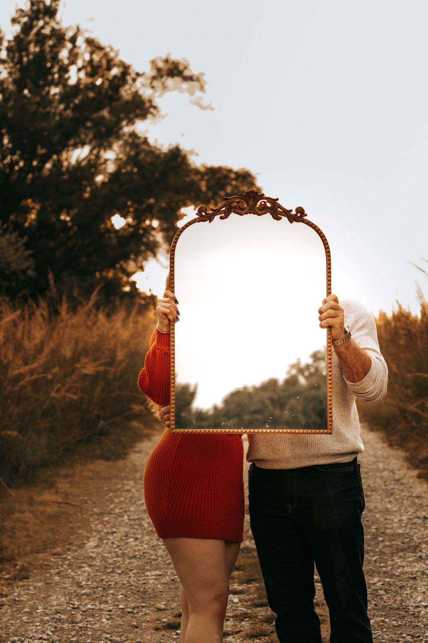 Two people hold an ornate vintage mirror on a dirt path at sunset creating an artistic optical illusion.