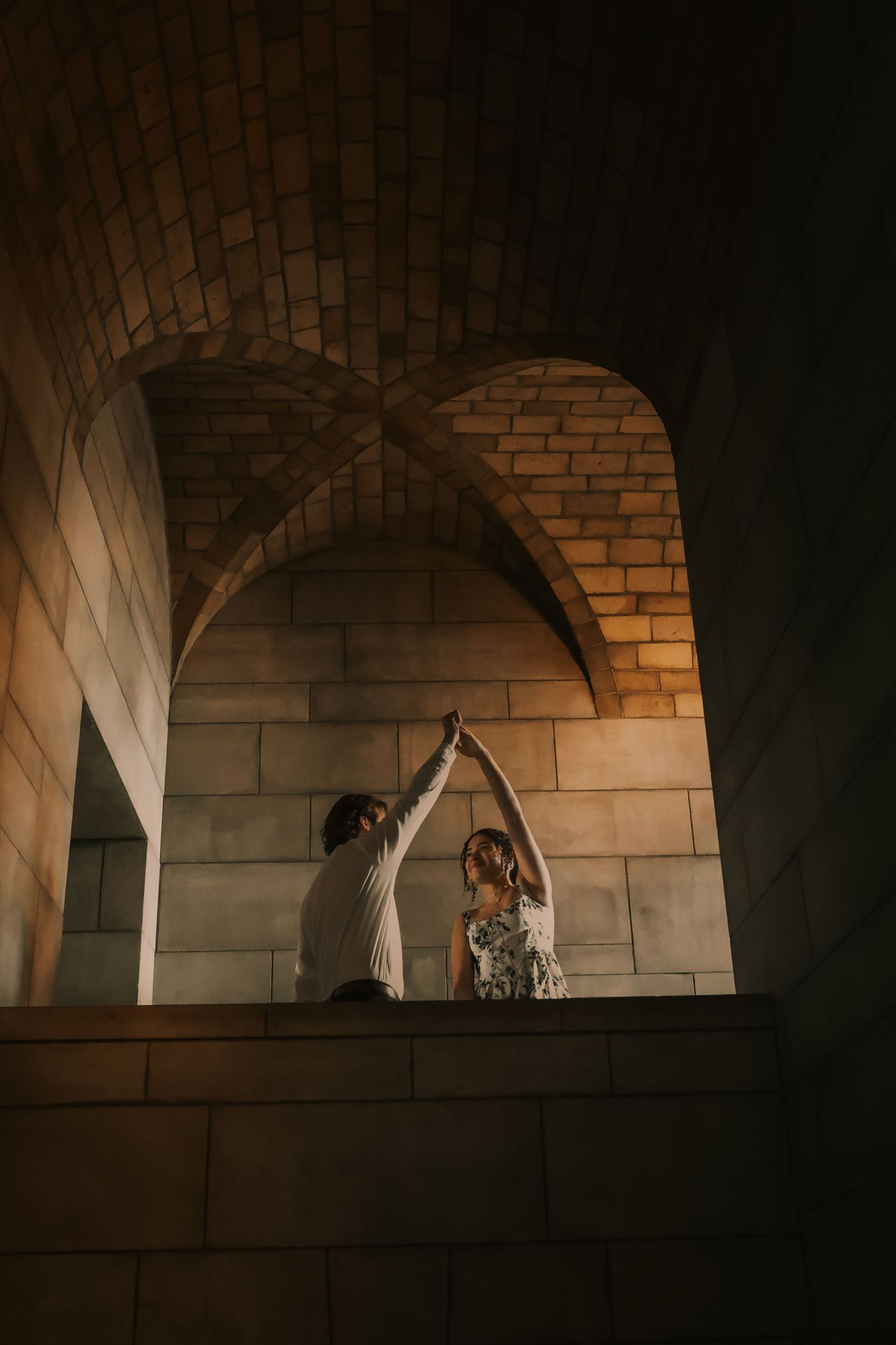 Two figures dance together under dramatic stone archways illuminated by warm lighting in an architectural setting.