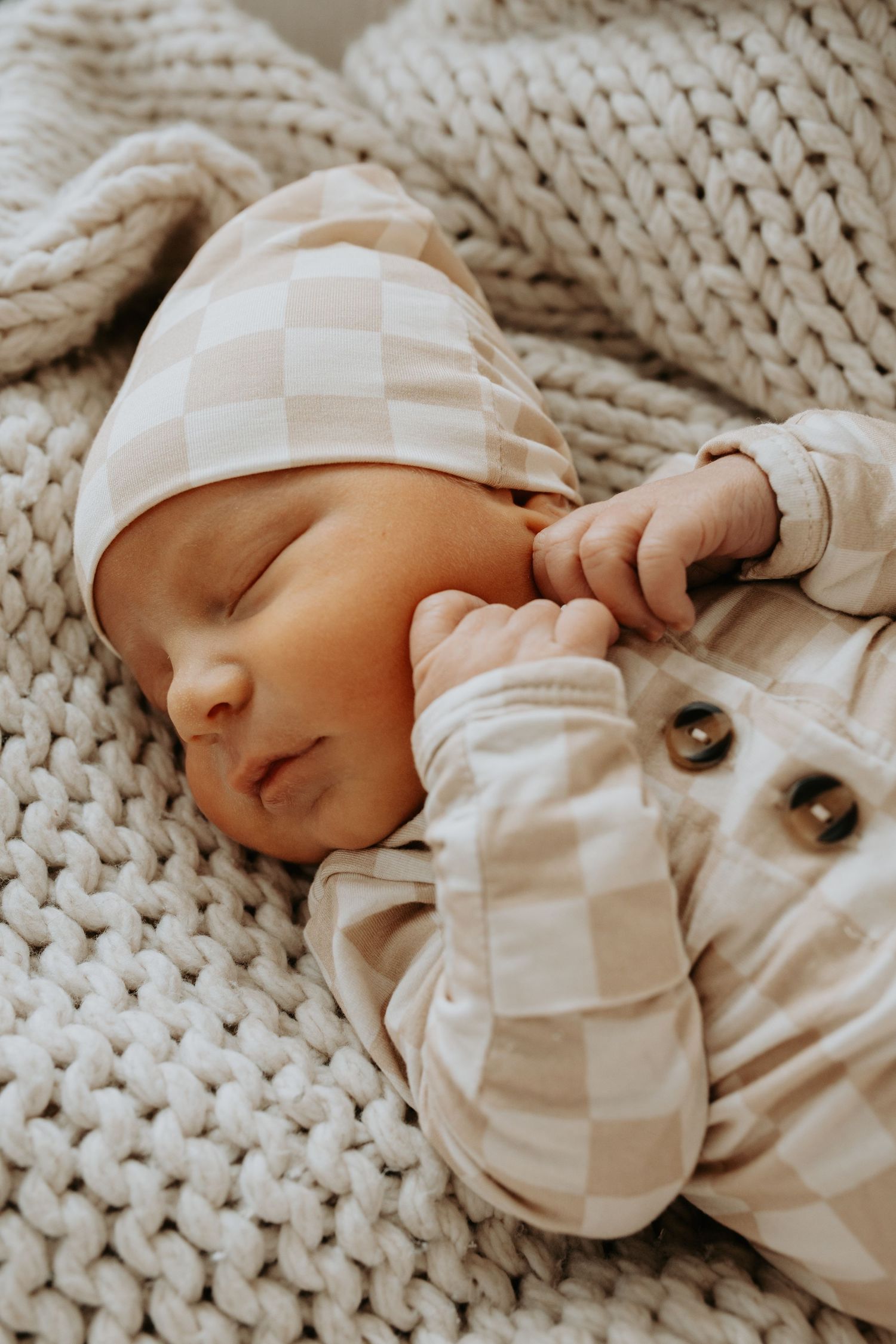 A sleeping newborn in a white striped outfit lies on a chunky knit cream-colored blanket.