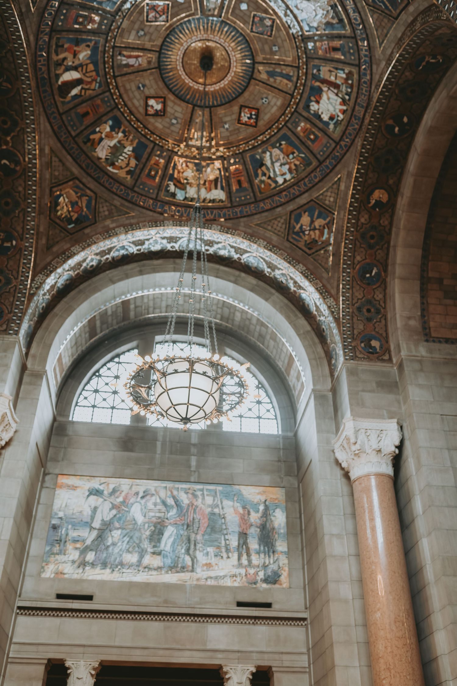 Ornate ceiling with intricate architectural details and murals in a grand historic building featuring arched windows and columns.