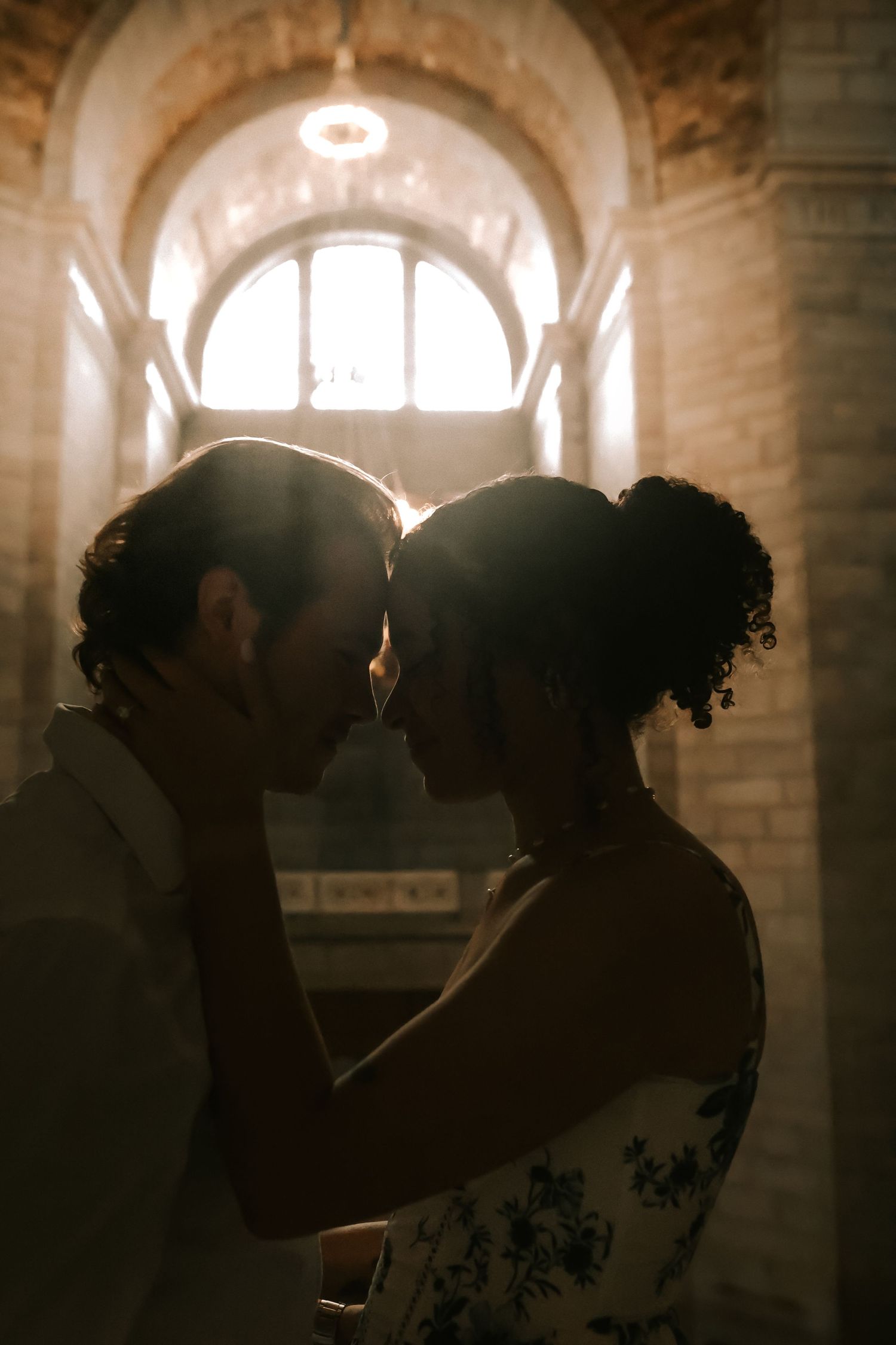 A romantic silhouette of a couple embracing in front of an arched window with beautiful backlighting in a historic building.