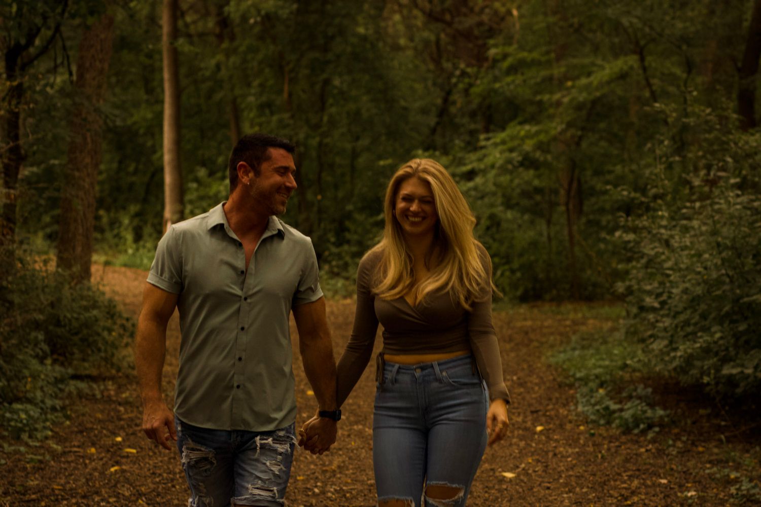 A couple walks hand in hand down a wooded path through a lush green forest during golden hour.