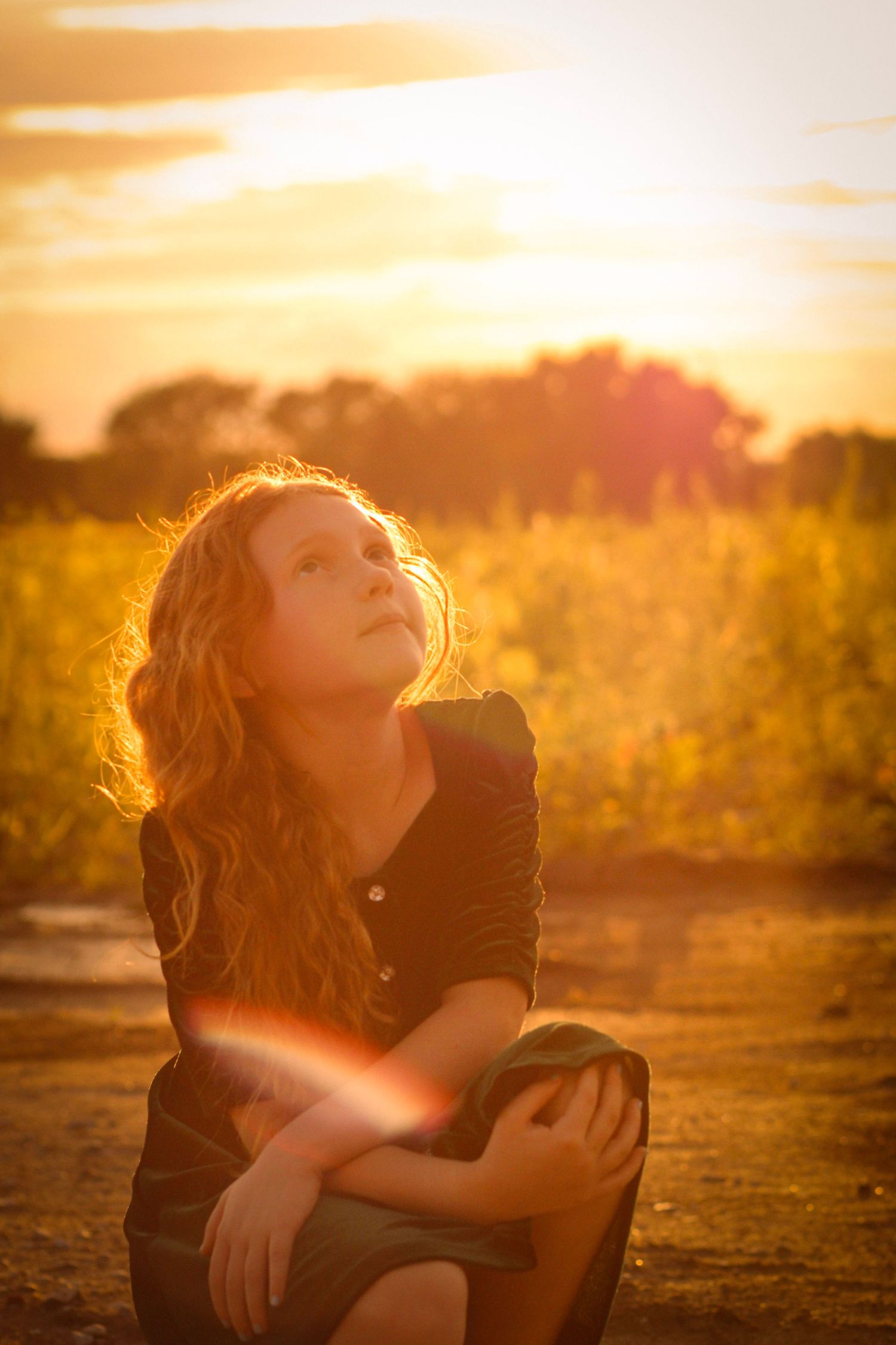 A person in a black outfit sits on the ground during golden hour as sunlight creates a warm atmospheric glow.