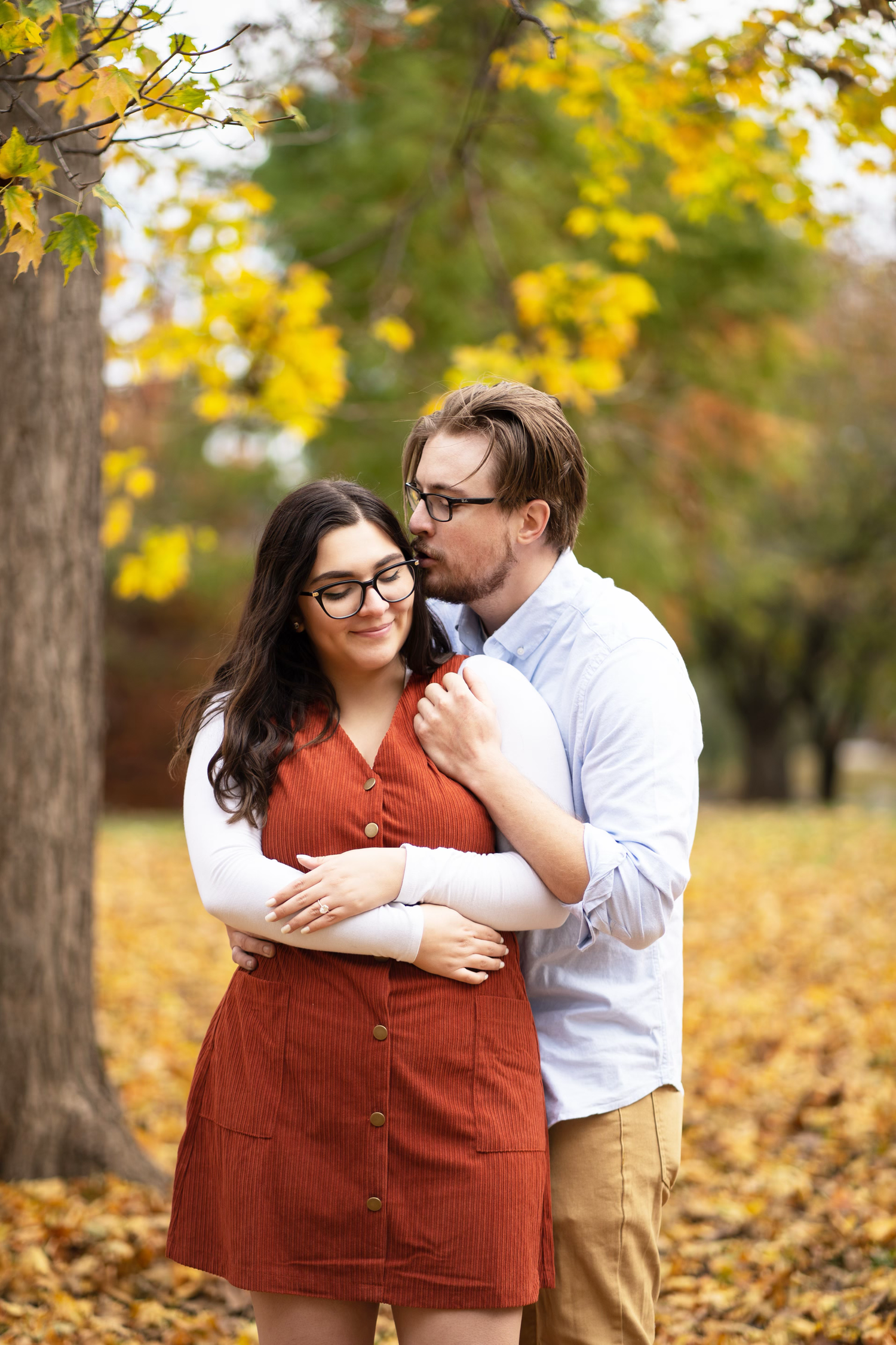 An engaged couple embraces for a photo among fallen autumn leaves in a park.