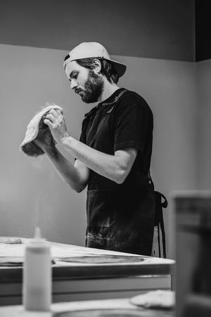 Panettiere con camicia e berretto neri che impasta la pasta su un tavolo da lavoro in bianco e nero.