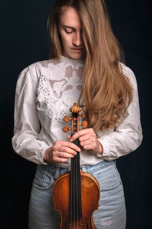 Una musicista con una camicetta di pizzo bianco e jeans tiene in mano un violino mentre guarda verso il basso su uno sfondo scuro.