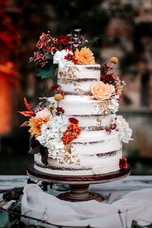 Una torta nuziale decorata con fiori bordeaux e pesca è appoggiata su un'alzata scura su uno sfondo sfocato.