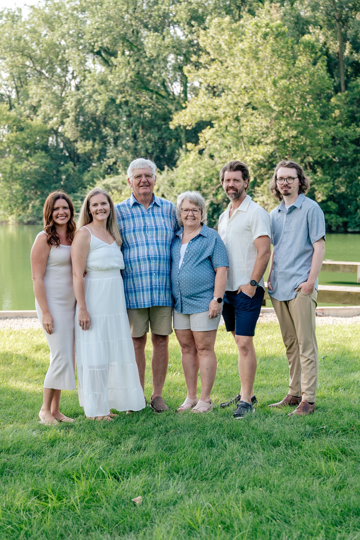 A group wearing coordinated neutral-toned outfits poses together outdoors on a sunny summer day near a lake.
