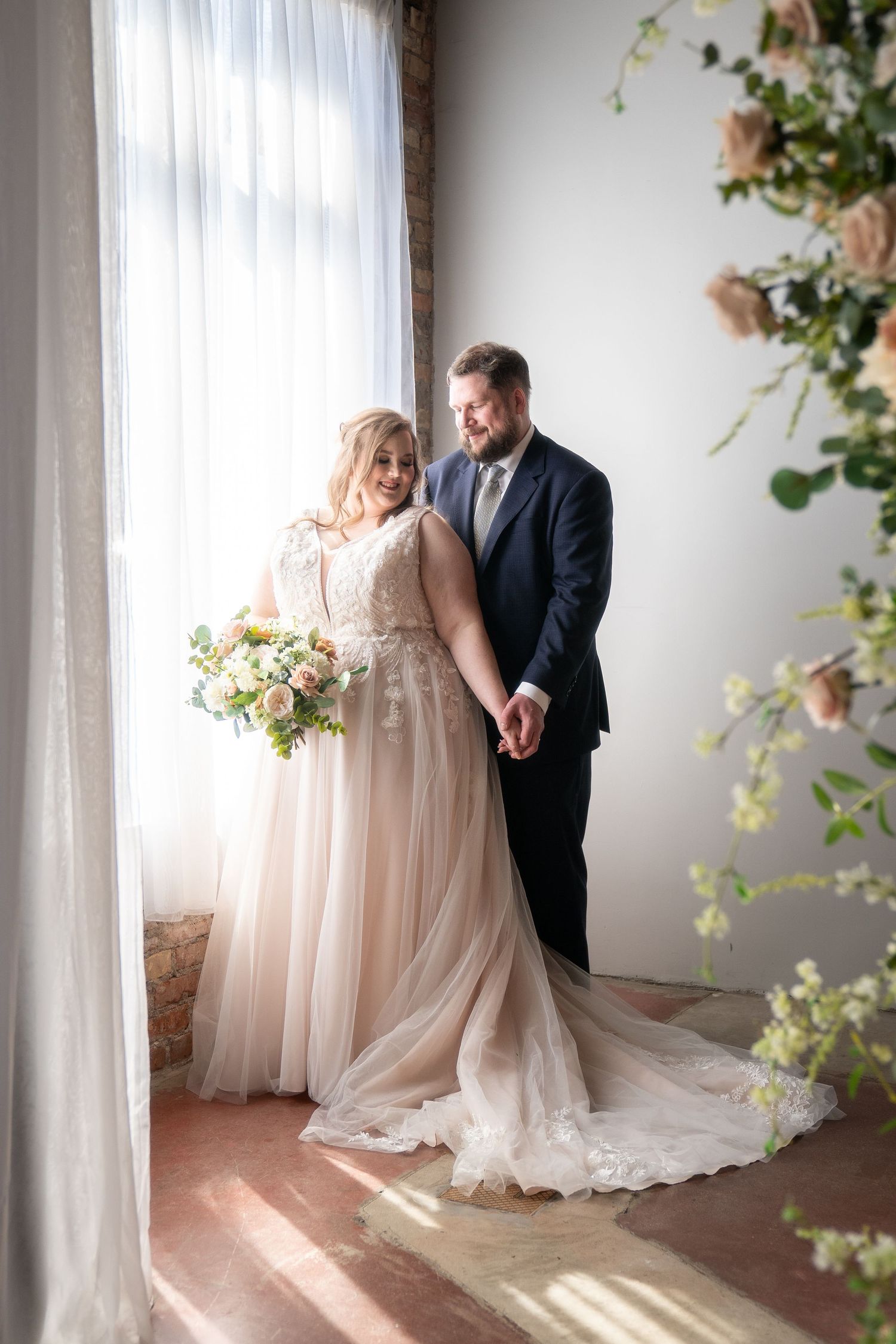 A romantic wedding couple poses together in a light-filled room with delicate floral decor and sheer curtains.