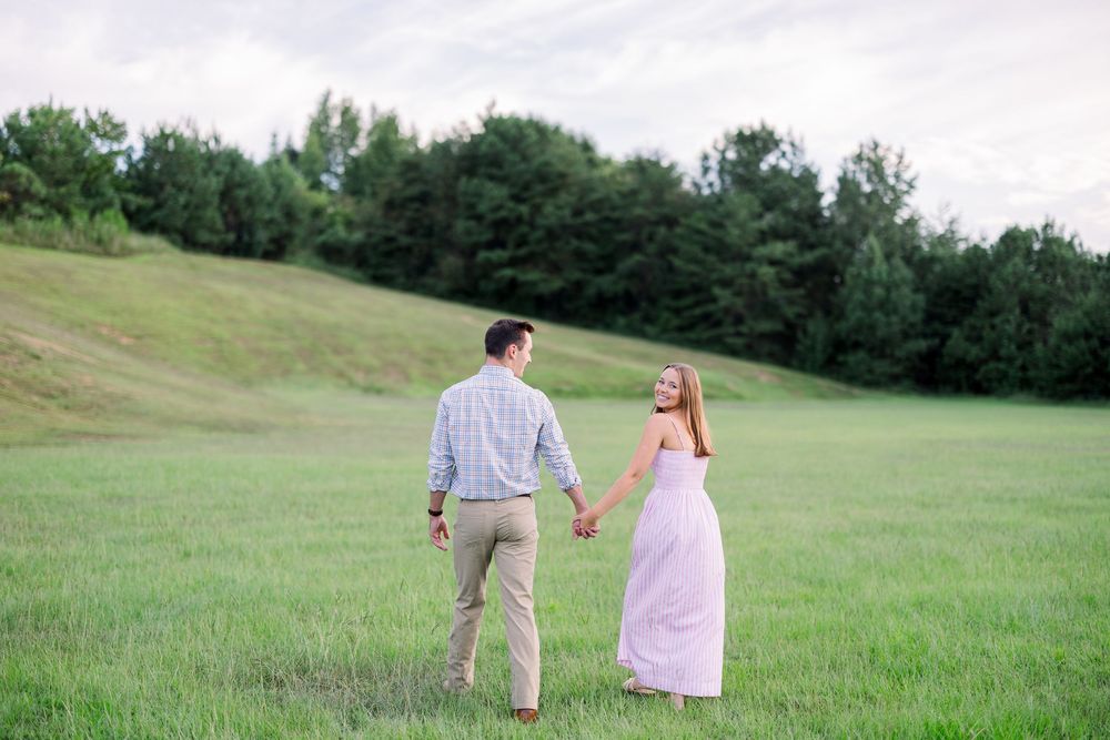 A couple dances playfully in a grassy meadow at sunset with trees in the background.