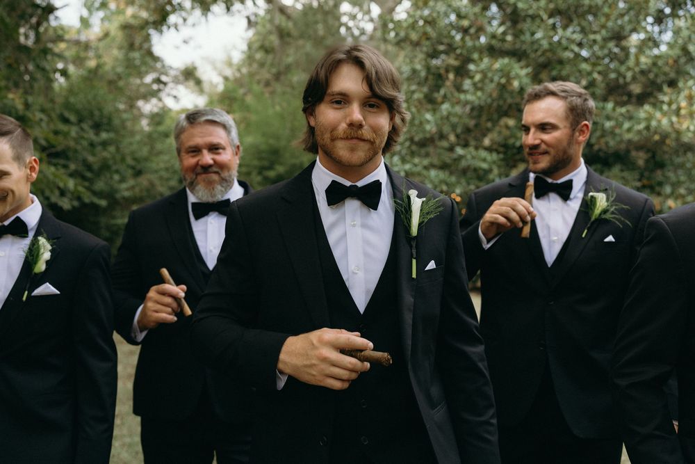 Groomsmen in black tuxedos and bow ties smile while walking together outdoors during a wedding celebration at The Elms of Coosada in Alabama