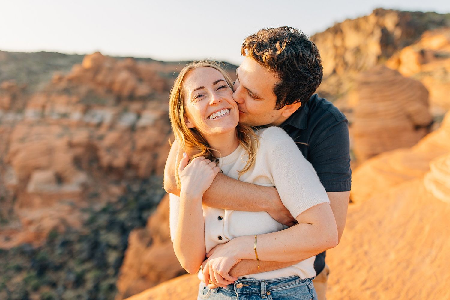 Couple embraces during sunset photoshoot in red rock desert landscape with warm golden light.