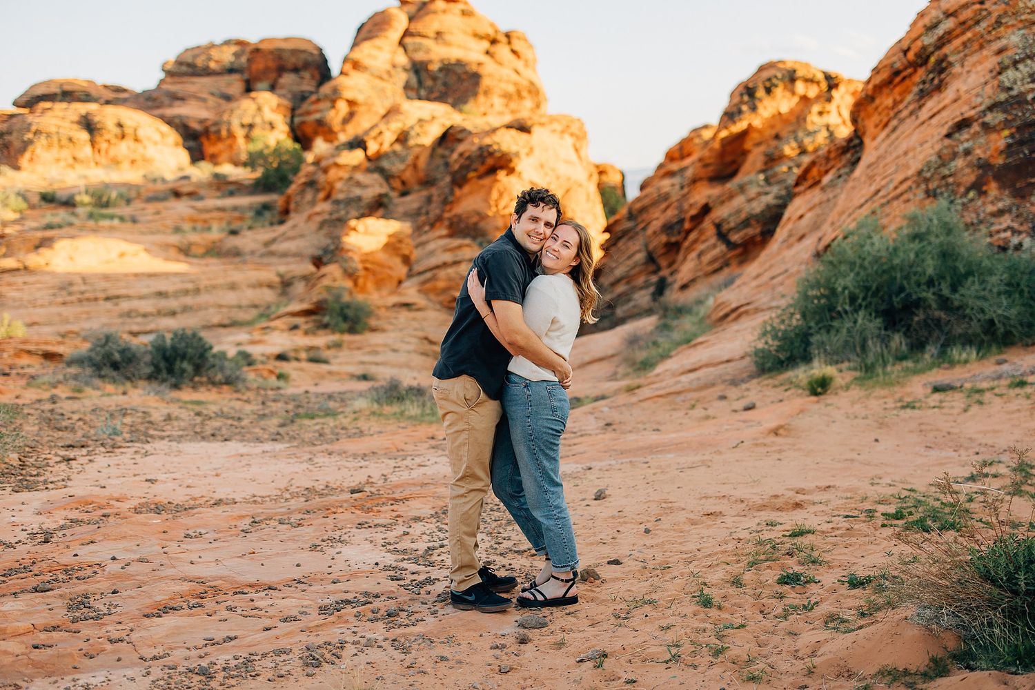 A couple embraces in front of stunning red rock formations at sunset in a desert landscape.
