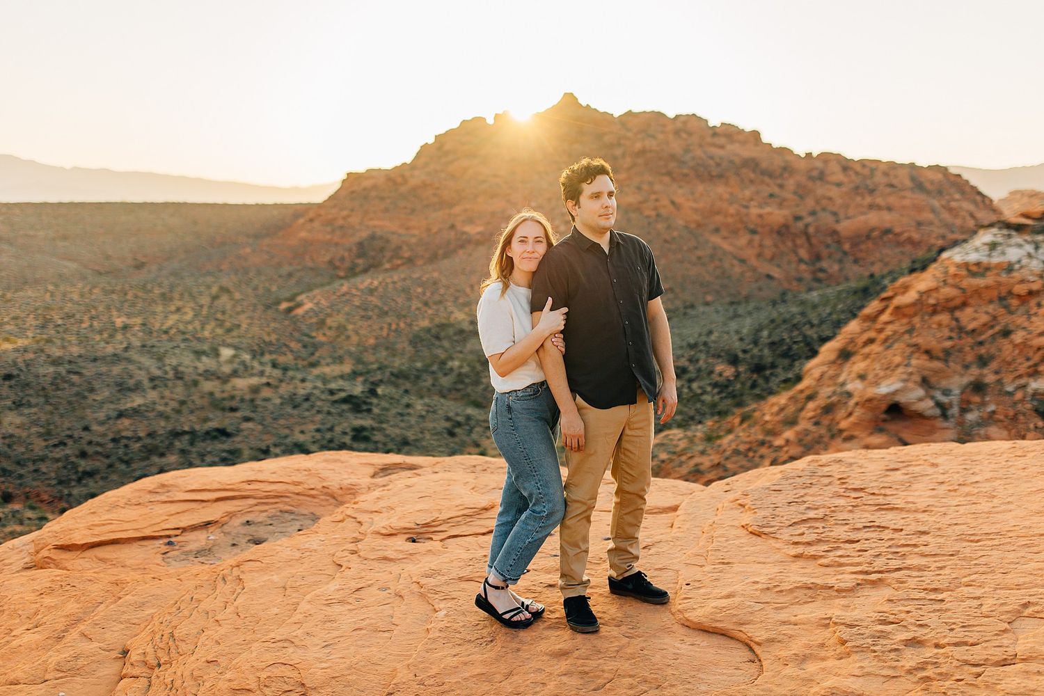 A couple embraces on red rock formations during golden hour at a scenic desert overlook in Utah.