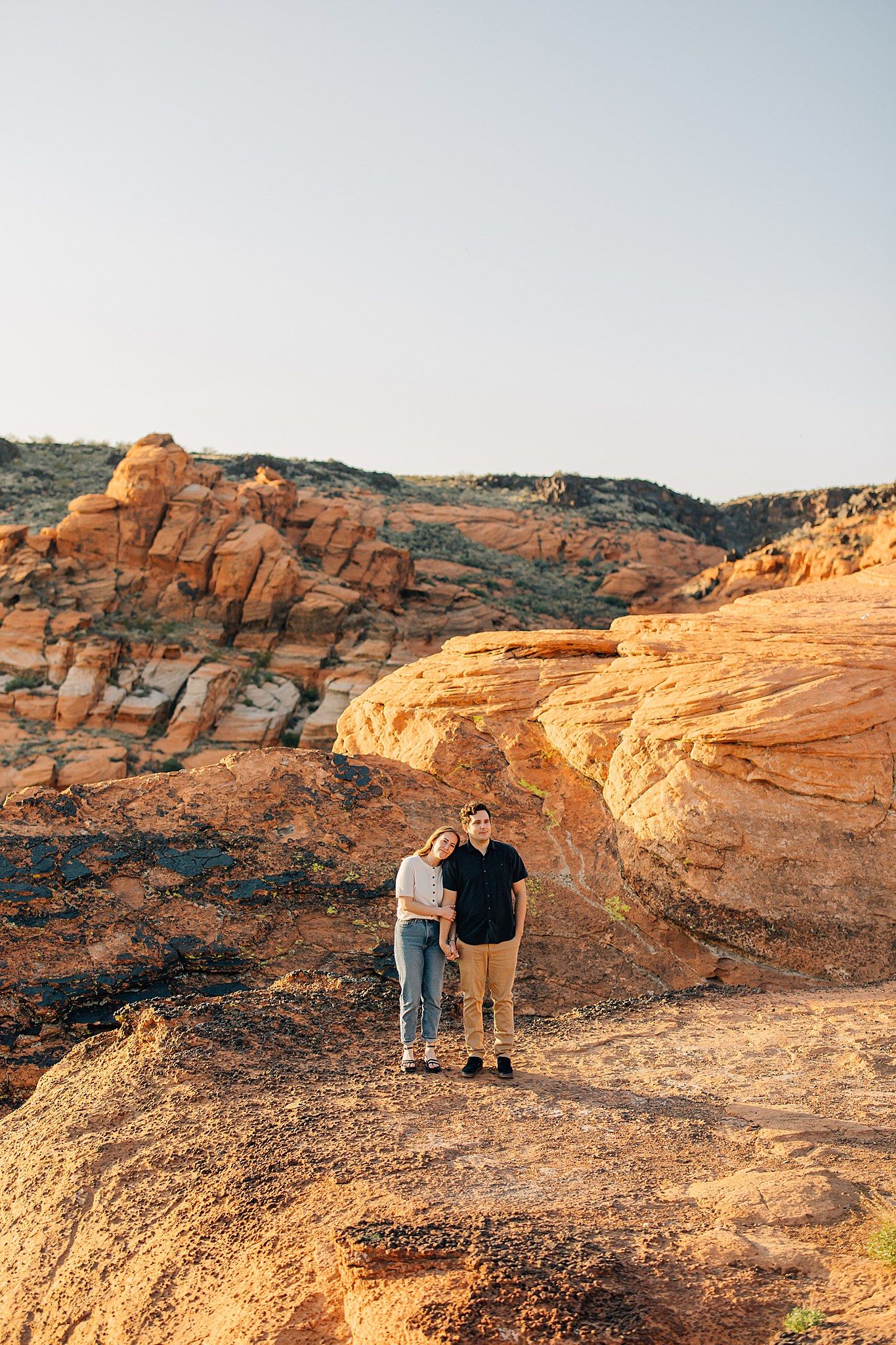 Two people stand together on red sandstone formations at sunset in a desert landscape.