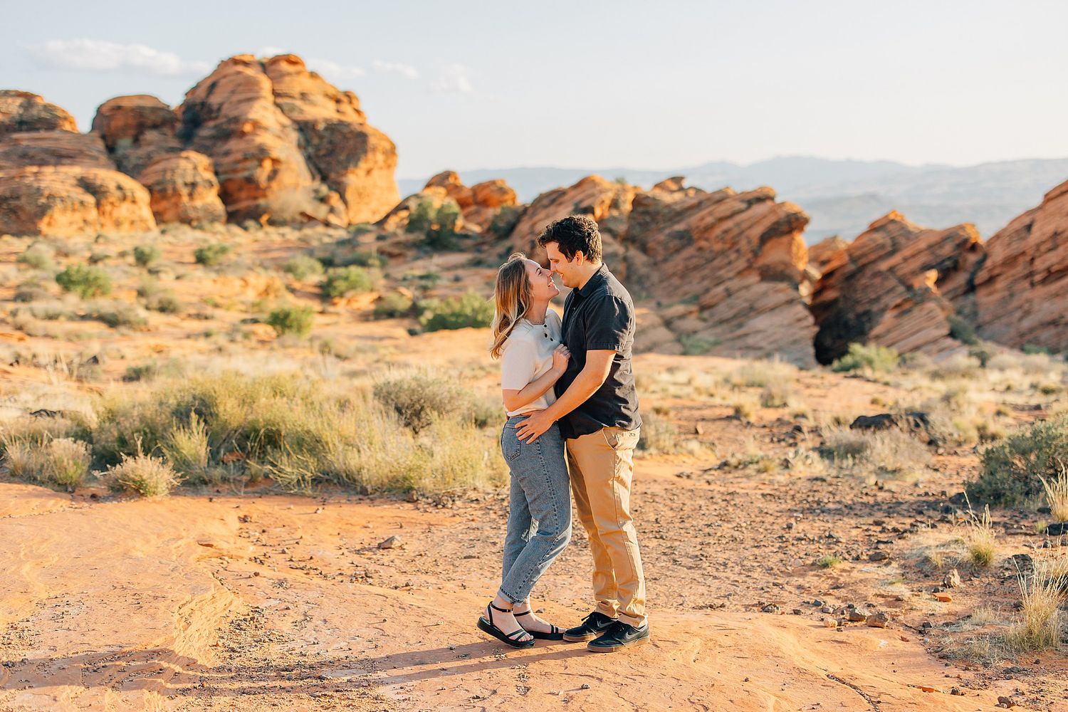 A couple embraces while exploring the scenic red rock formations in a desert landscape at sunset.
