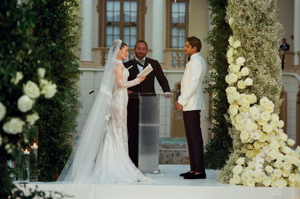 A romantic wedding ceremony moment captured in front of a grand venue with elegant white floral arrangements.