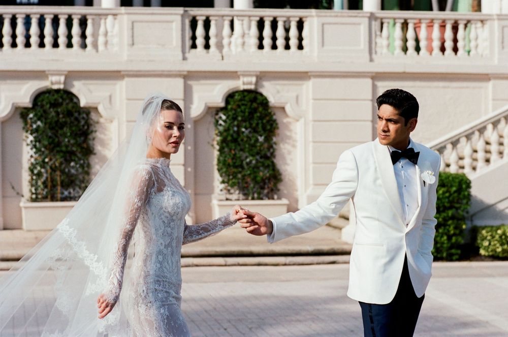 Bride in lace gown with veil holds groom's hand in white tuxedo jacket outdoors at elegant venue.