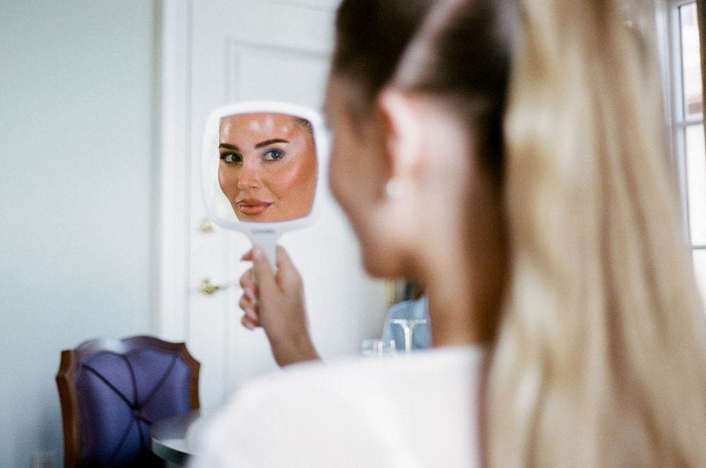 A bride in a white wedding dress gets ready with her makeup and hair being styled in a bright room.