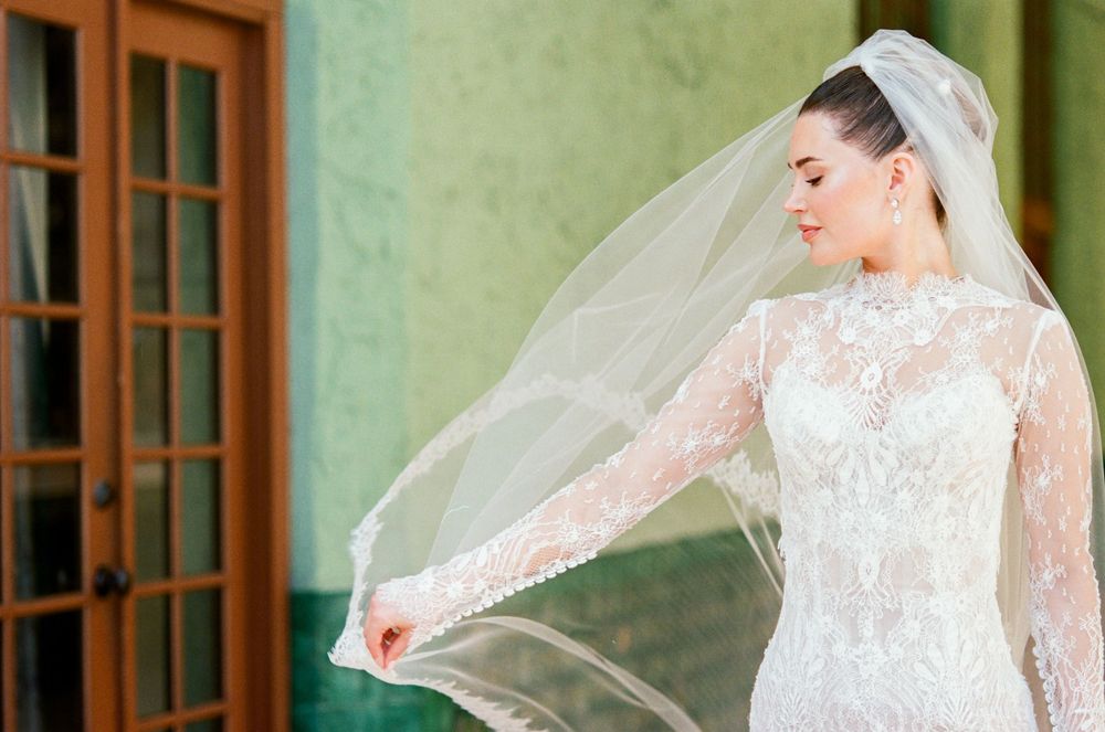 A bride in an elegant lace wedding dress and veil stands in soft lighting near windows.
