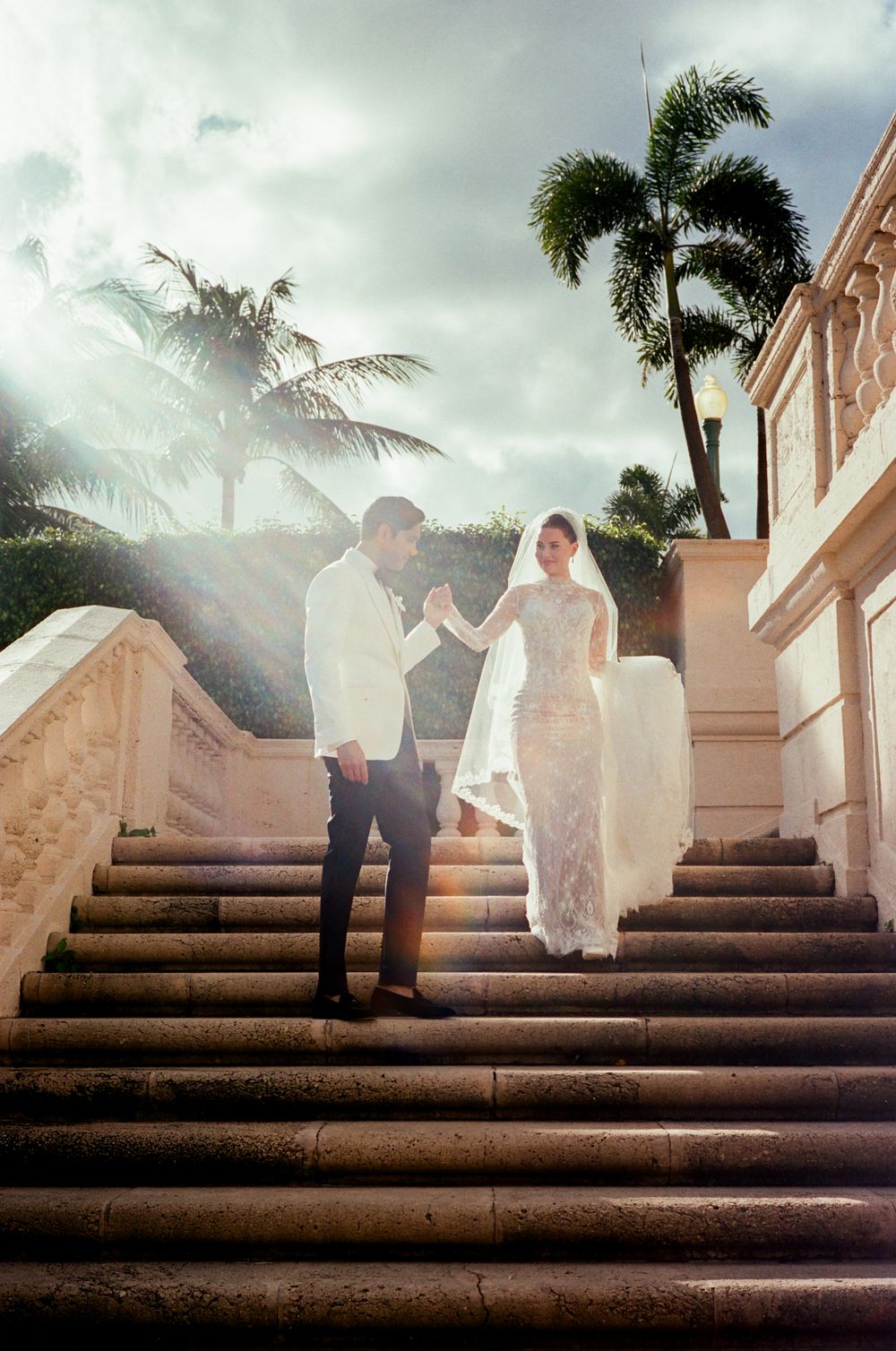 A romantic wedding couple ascends stone steps in a tropical setting with dramatic moody skies and palm trees.