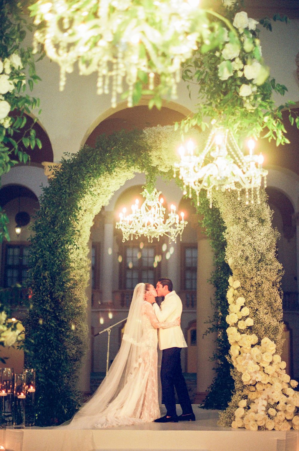Bride and groom share a kiss under an elegant floral arch adorned with greenery and chandeliers at a romantic wedding.