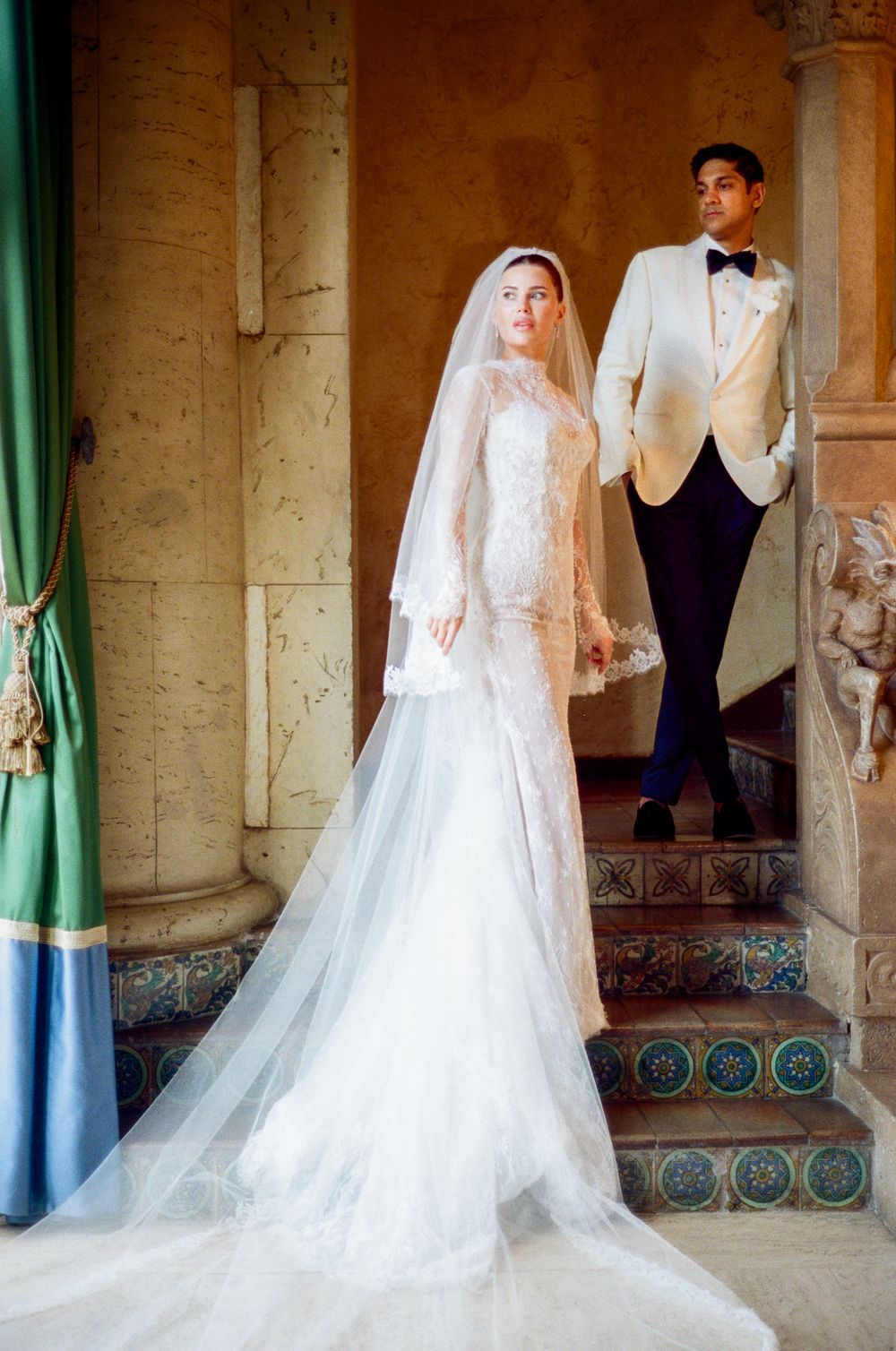 A bride and groom pose in an elegant colonnade with Spanish colonial architecture and green curtains.