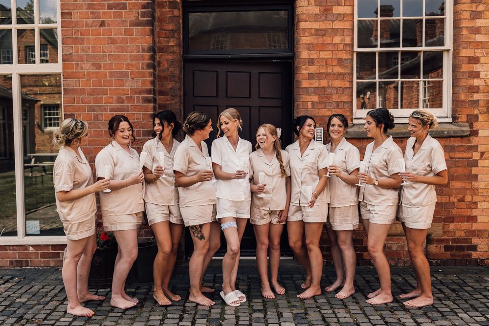 Wedding party members in light colored dresses posing in a line against a brick wall exterior.