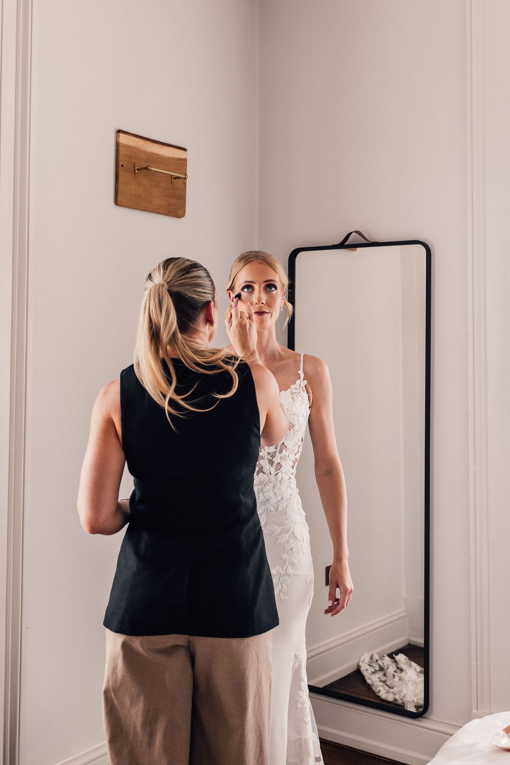 Woman in black top helps another try on a white wedding dress in front of a mirror.