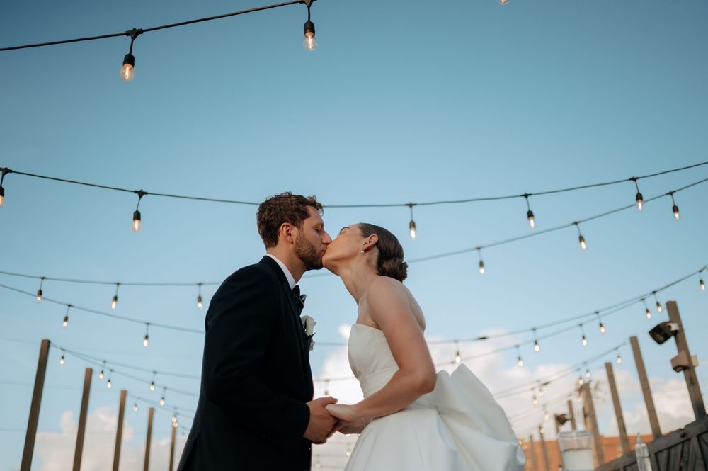 Bride and groom share a kiss under string lights on a rooftop at dusk.