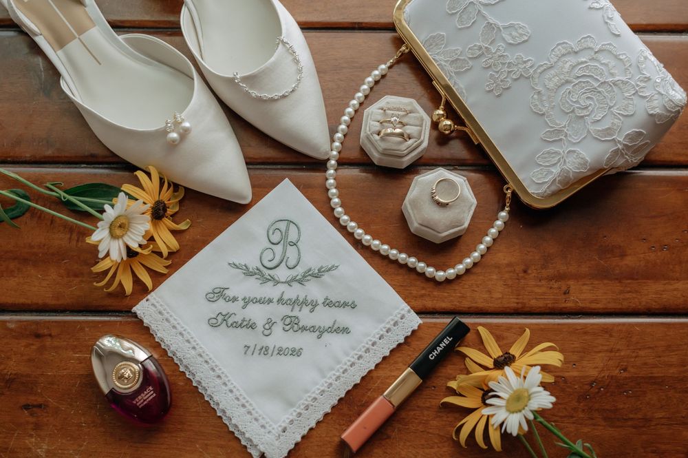 Wedding flat lay with embroidered handkerchief, pearl necklace, white heels, and daisy flowers.