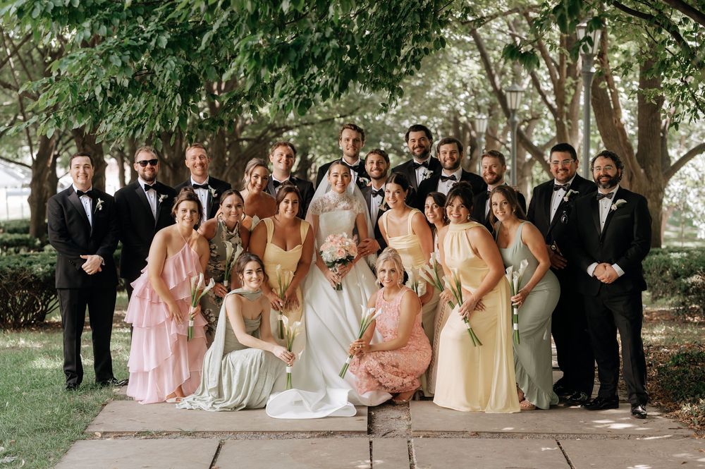 Large wedding party poses together outdoors under trees, bridesmaids in pastel dresses and groomsmen in black tuxedos.