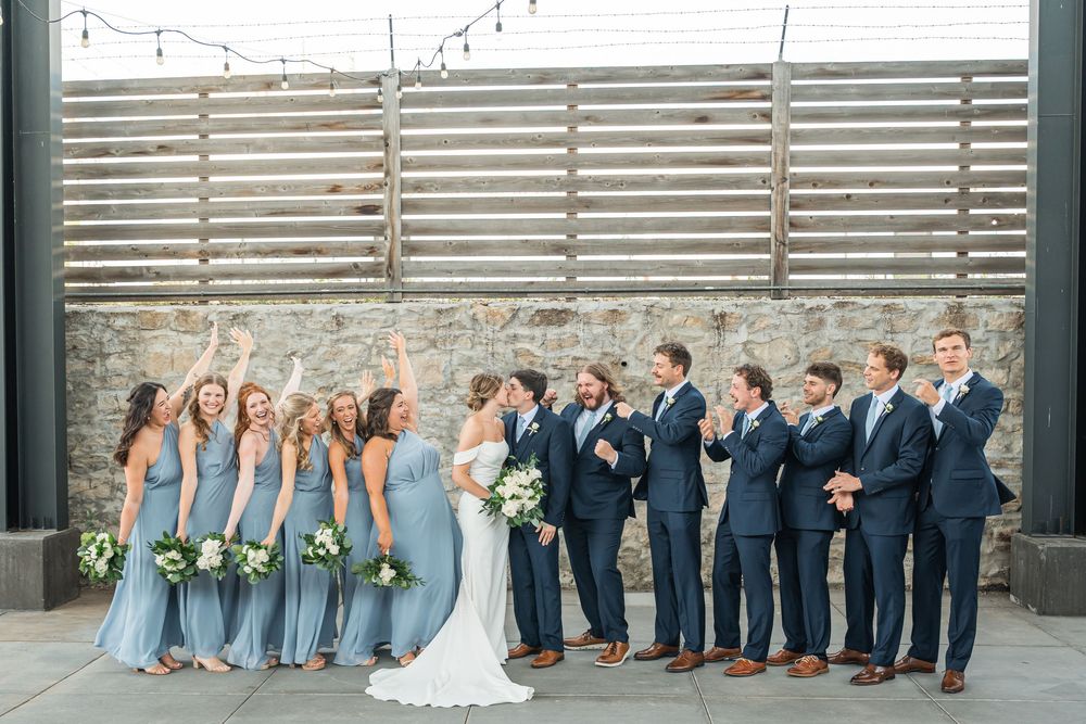 Wedding party poses together, bridesmaids in dusty blue gowns and groomsmen in navy suits, celebrating outdoors near stone wall.