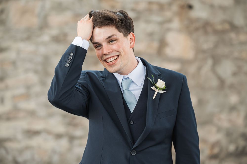 Smiling groom in navy suit with boutonniere poses playfully against stone wall at outdoor wedding venue.