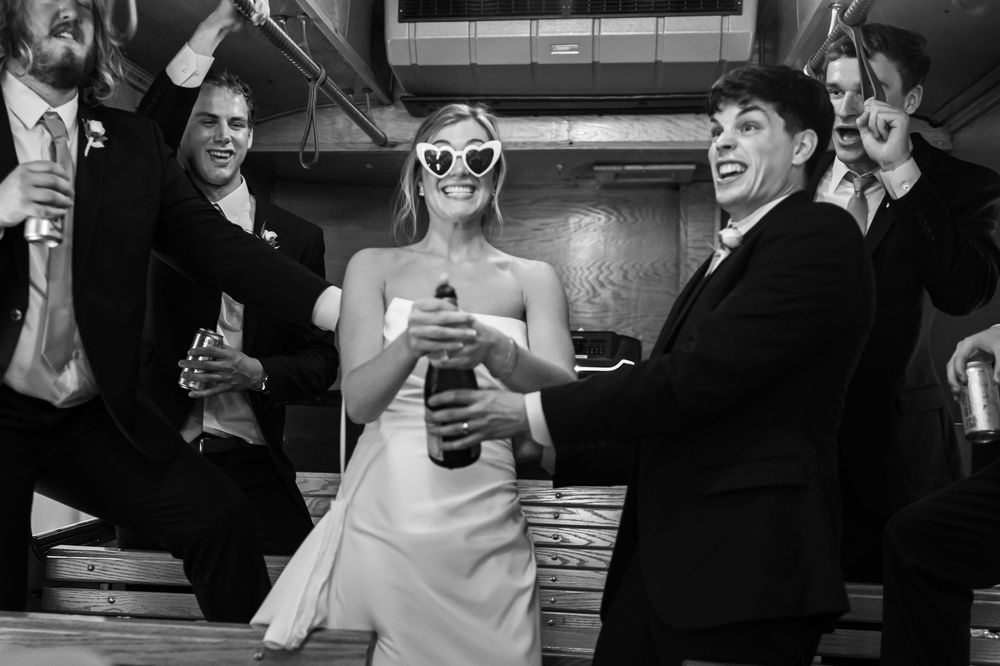 Bride wearing heart sunglasses opens champagne bottle with groom and wedding party in black and white photo.