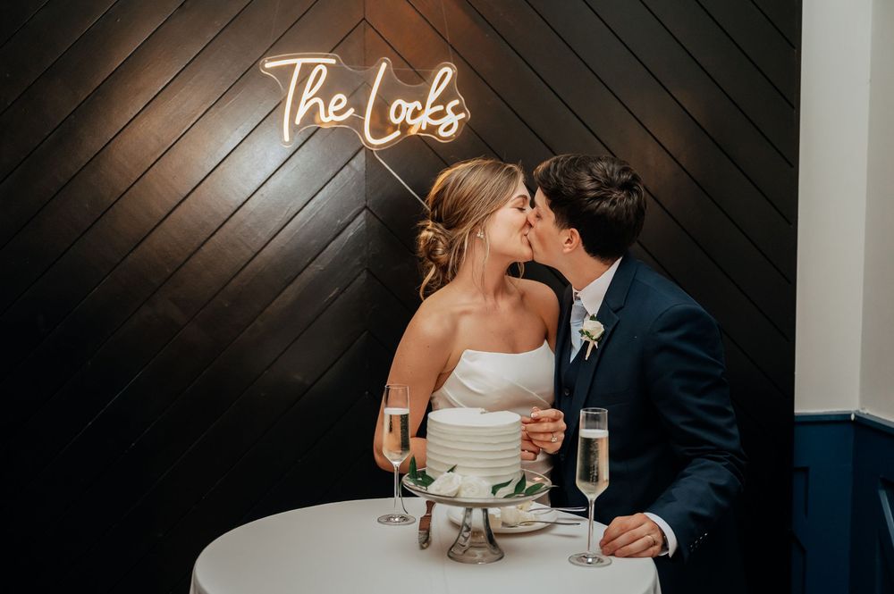 Newlyweds kiss at cake table beneath glowing neon sign reading The Locks at wedding reception.