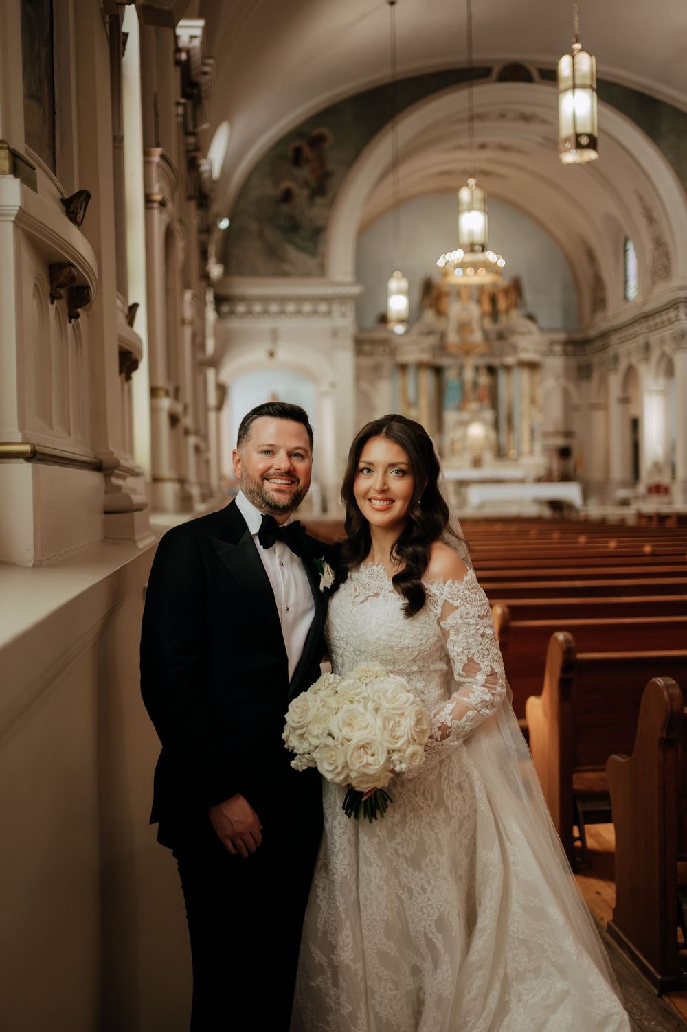Bride in lace off-shoulder gown and groom in black tuxedo pose together inside an elegant church.